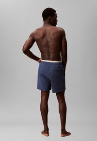 Costume da bagno blu navy con vita elasticizzata, dettaglio della tasca posteriore e una texture del tessuto liscia. Modello mostrato di schiena.