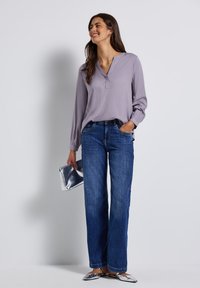 Blusa grigia chiaro a maniche lunghe con scollatura a V, abbinata a jeans larghi blu. Tiene una pochette argento e indossa scarpe slip-on argentate.