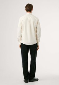 Crèmegekleurde shirt met knoopmanchetten, eenvoudige uitstraling en rugpasdetail, gecombineerd met donkerblauwe broek en zwarte schoenen.