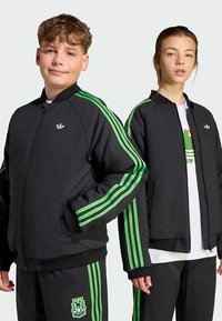 Black athletic jackets featuring green stripes, zip front, and side pockets, worn with coordinating pants. Logo displayed on the left chest.