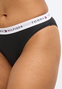 Perizoma nero con vita elastica bianca stampata con "TOMMY HILFIGER." Tessuto liscio, design aderente e cuciture minime.