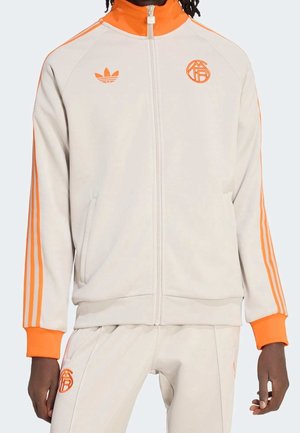 Personne portant un survêtement beige avec des bandes orange, col orange, poignets et logos assortis sur la poitrine et la cuisse.