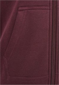 Gros plan sur une poche en tissu bordeaux avec des coutures visibles et une fermeture éclair métallique partiellement fermée sur le côté droit.