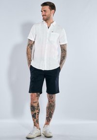 Kurzärmliges weißes Hemd mit Knopfleiste und strukturiertem Muster, Brusttasche und marineblauen Shorts. Das Modell trägt weiße Sneaker und zeigt Tattoos an den Beinen.
