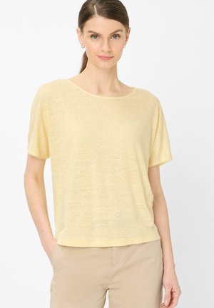 Frau mit braunen Haaren in einem Pferdeschwanz, die ein hellgelbes kurzärmeliges Oberteil und beige Hose trägt, steht mit einer Hand in der Tasche.