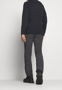 Pull en maille noir pour homme avec des poignets côtelés, associé à un pantalon gris, fuselé et isolé, et des chaussures noires avec texture.