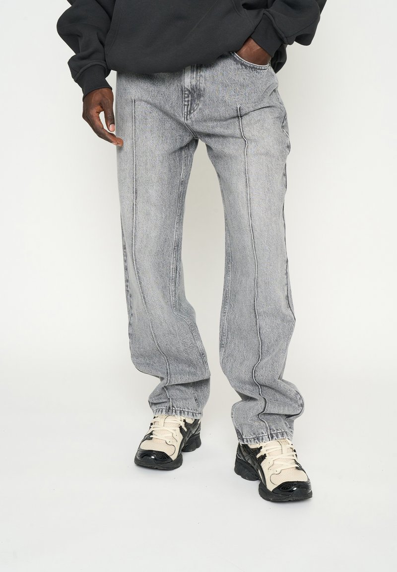 Jeans de mezclilla gris con un corte holgado, con bolsillos frontales y detalles de dobladillo desgastado, combinados con zapatillas gruesas en negro y crema.