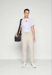 Biely skrátený tričko s čiernym logom, béžové zúžené nohavice a biele tenisky. Čierna taška s ramenným popruhom držaná v ľavej ruke.