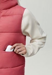 Vadderad, rosa ärmlös väst med hög krage och en vit långärmad skjorta under. Hand som lägger ett vitt föremål i en ficka.