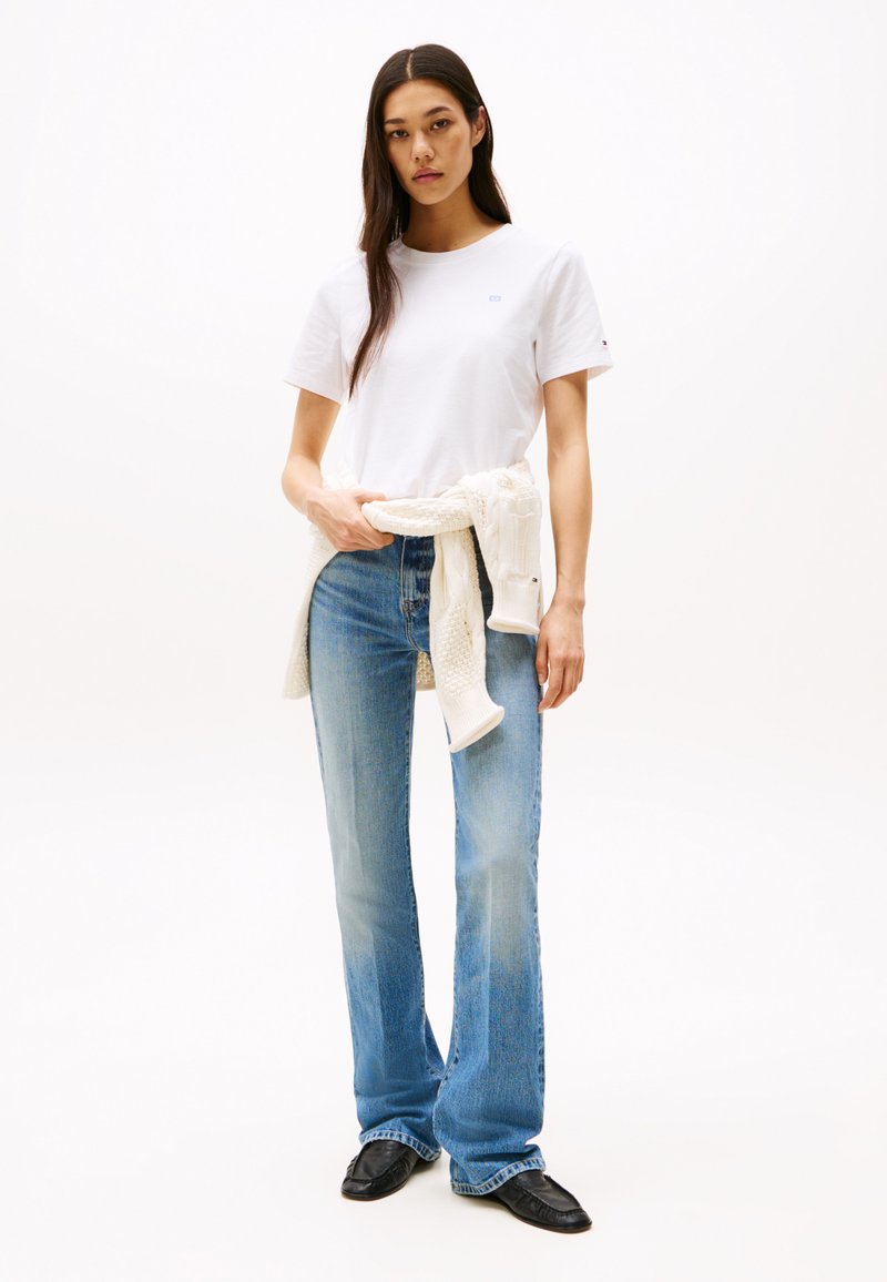 Camiseta blanca de manga corta con logotipo azul, pantalones vaqueros rectos de un azul claro y zapatos de cuero negros. Suéter crema atado a la cintura.