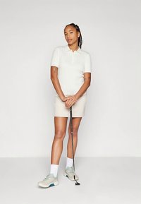 Mulher com cabelo trançado, vestindo uma polo de malha branca, saia bege, meias e sapatilhas, segurando um taco de golfe, em pé contra um fundo simples.