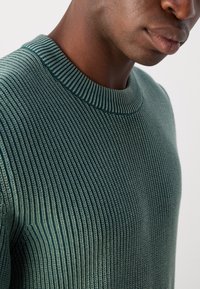 Pull en tricot vert avec une texture côtelée, un col rond ajusté et des motifs à rayures verticales. Le matériau semble léger et doux.