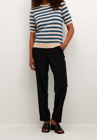 Kobieta ubrana w niebiesko-beżowy paskowany sweter z krótkim rękawem, czarne spodnie i czarne sandały na obcasie, stojąca z jedną ręką w kieszeni.