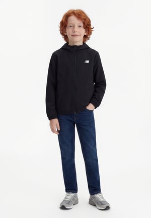 Ung rødbrun dreng står med hånden i lommen iført sort hættetrøje med lynlås, blå jeans og grå New Balance-sneakers på hvid baggrund.