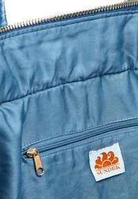 Tote en tissu bleu avec une poche zippée dotée d'une fermeture éclair dorée. Une étiquette blanche affiche un logo de soleil orange avec le texte "SUNDEK". Texture lisse.