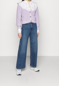 Ljuslila stickad kofta med en stor vit ögla spetskrage, kombinerad med högmidjade blå vida jeans och vita sneakers.