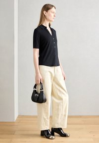 Camicia nera a maniche corte con bottoni, pantaloni beige a gamba larga, borsa nera, e stivaletti neri in pelle verniciata con punta metallica.