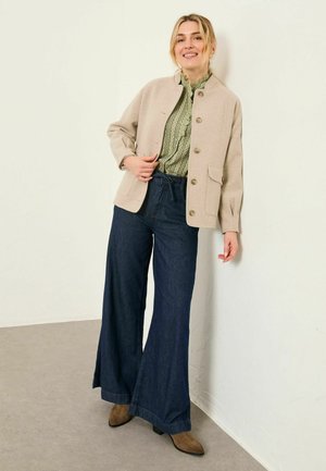 Beige Jacke aus Wollmischung mit Knopfverschluss und Taschen, getragen über einer grünen, gemusterten Bluse und dunklen Jeanshosen mit weitem Bein. Braune Stiefeletten.