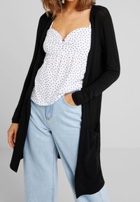 Black long cardigan made of soft fabric with open front and side pockets, worn over a white polka dot top and light blue jeans.