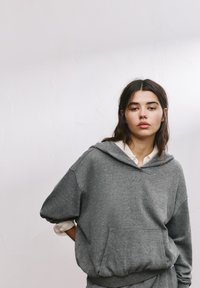 Hanorac gri cu glugă, realizat dintr-un material moale, cu o buzănele frontală și tiv elastic. Croială lejeră cu umeri lăsați.