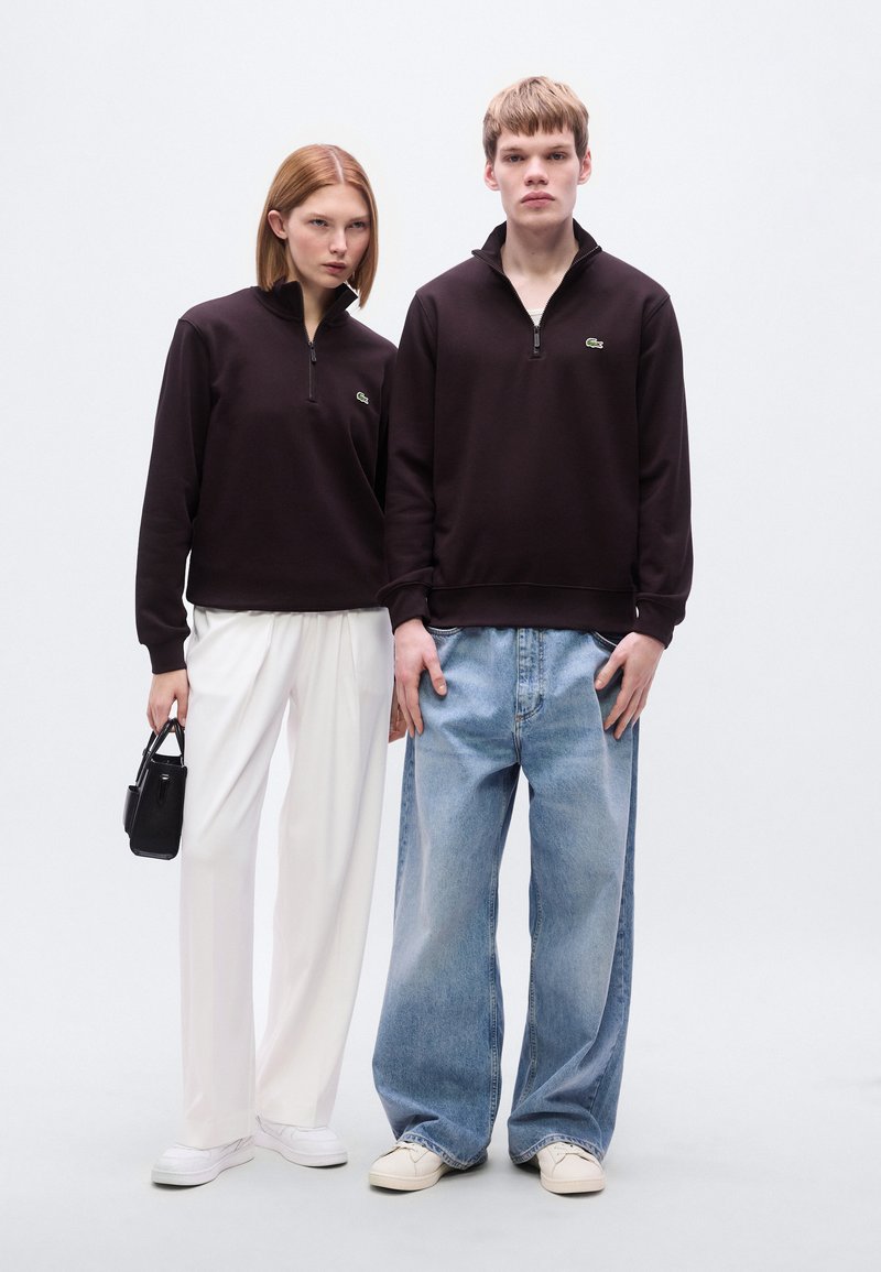 Des sweats à capuche noirs zippés portés par deux personnes, l’une en pantalon blanc, l’autre en jean bleu ample, avec un petit logo en accent sur les sweats.