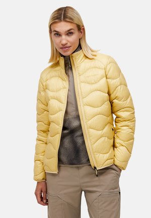 Kvinde med blondt hår iført en gul quiltet jakke, gråt inderlag og beige bukser med hænderne i lommerne mod en ensfarvet baggrund.