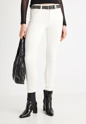 Vaqueros blancos de corte ajustado, combinados con un cinturón negro con tachuelas y botines negros brillantes. Se sostiene un bolso de mano negro con textura.