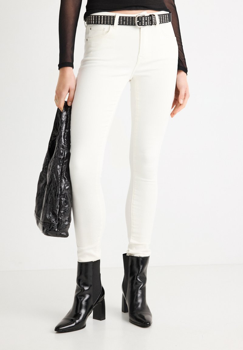 Λευκό στενό τζιν με εφαρμοστή σχεδίαση, συνδυασμένο με μαύρη ζώνη με καρφιά και λαμπερά μαύρα μποτάκια στον αστράγαλο. Κρατιέται υφασμάτινη μαύρη τσάντα με υφή.