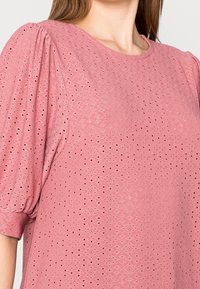 Valkoisen tekstuurin ja timanttikuvion omaava vaaleanpunainen puffihihainen paita, jossa on pyöristetty pääntie ja pehmeä laskos.