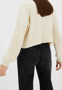 Vrouw in een crèmekleurige ribgebreide cropped sweater met ballonmouwen en hoge taille zwarte jeans, gezien van achteren.