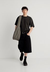 Νεαρό άτομο περπατά φορώντας μαύρα μεγάλα σορτς, μαύρο μπλουζάκι, μαύρα παπούτσια και μια μεγάλη γκρι τσάντα ώμου σε λευκό φόντο.