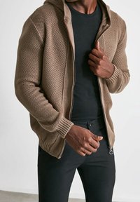 Felpa a zip marrone in maglia con motivo testurizzato, cappuccio foderato e polsini a coste, indossata sopra una maglietta nera e abbinata a pantaloni scuri.