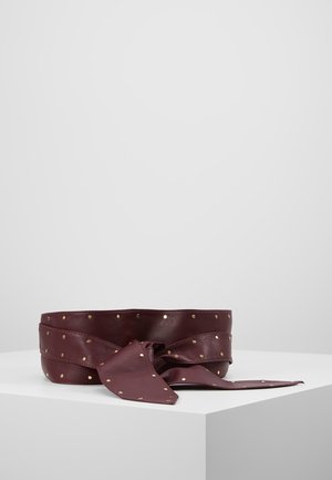 Donkerrode leren riem met studs in goudkleur, geknoopt en getoond op een wit voetstuk tegen een effen grijze achtergrond.