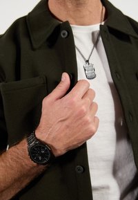 Schwarze Uhr mit runder Ziffernblatt und Metallarmband, übereinander getragen auf einem weißen T-Shirt, Hände auf einer dunkelgrünen Hemdbluse, silberne Hundemarke als Halskette.