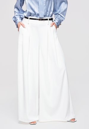 Kvinde iført hvide bukser med høj talje og vide ben med sort bælte, lyseblå satinbluse, hænder i lommerne og firkantede hælede sandaler.