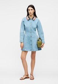 Γυναίκα με μακριά σκούρα μαλλιά που φοράει ανοιχτό μπλε φόρεμα από τζιν με πολύχρωμο γιακά, κρατώντας μια μικρή τσάντα σε ελιά πράσινο και φορώντας καφέ σανδάλια.