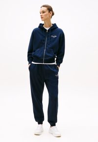Sweat à capuche zippé bleu marine avec cordons, poches avant et logo. Associé à un pantalon de jogging assorti et des baskets blanches, avec des poignets côtelés.