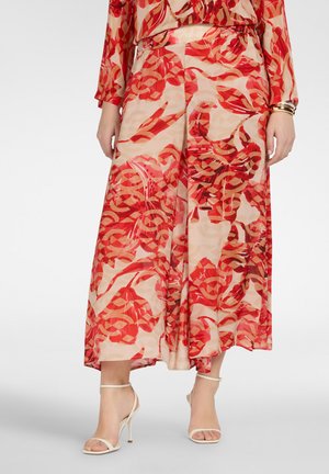 Frau trägt einen fließenden Rock und ein Oberteil mit rotem und beigem abstraktem Blumenmuster, kombiniert mit weißen High-Heel-Sandalen und goldenen Armbändern.