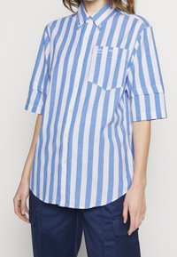 Persona che indossa una camicia a maniche corte azzurra e bianca a righe verticali con taschino sul petto e pantaloni cargo blu scuro.