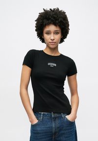 Czarny prążkowany t-shirt z krótkimi rękawami, z okrągłym haftowanym logo „GUESS” na piersi. Zestawiony z niebieskimi dżinsami.