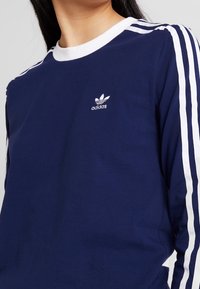 Laivastonsininen pitkähihaisella t-paidalla, jossa on valkoinen ribbauskaulus ja kolme valkoista raitaa jokaisessa hihassa. Paidassa on pieni valkoinen adidas-logo rinnassa.