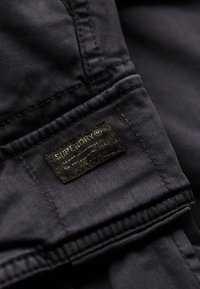 Donker denimstof met zichtbare stiksels, voorzien van een zwart leren label met het merk "Superdry" en aanvullende tekstdetails.