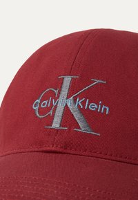 Rode katoenen pet met een zilveren 'CK'-logo en 'Calvin Klein' in blauwe borduursel, voorzien van een gestructureerde klep en gestikte panelen.
