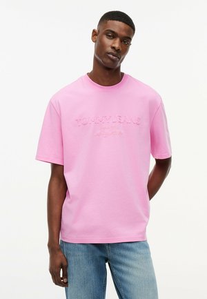 Man draagt een ruim vallend roze T-shirt met geborduurde tekst "Tommy Jeans" en lichtblauwe spijkerbroek, staand tegen een effen witte achtergrond.