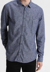 Blauwe button-up blouse gemaakt van textuurstof, met een klein wit patroon, een borstzak en lange mouwen. Halflange kraag.