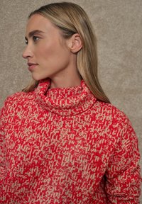 Jersey de cuello alto en rojo y crema con un patrón, que presenta un alto cuello y una tela texturizada, con un corte holgado y un diseño abstracto.