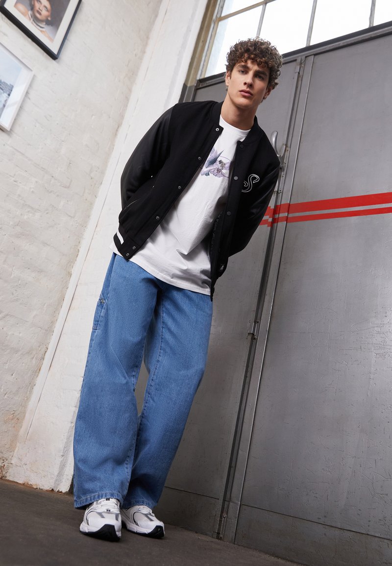 Sort varsity-jakke med læderærmer, hvid grafisk t-shirt, løse blå jeans og hvide sneakers, der står op ad en grå væg.