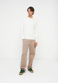 Ung mand står iført en langærmet hvid skjorte, beige bukser og grønne sneakers foran en ensfarvet hvid baggrund.