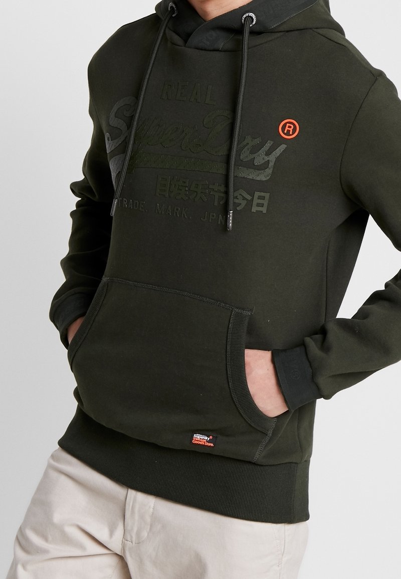 Personne portant un sweat à capuche vert foncé avec une poche avant et un logo Superdry discret, mains dans les poches, associée à un pantalon beige.