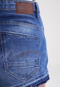 Jeansshorts i en blekblå färg, med fransig kant och en brun läderlogotyp vid midjan. Bakfickan har en karakteristisk söm.